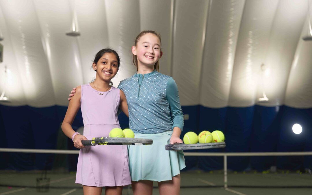 Two girls holding their rackets and smiling during junior tennis training.