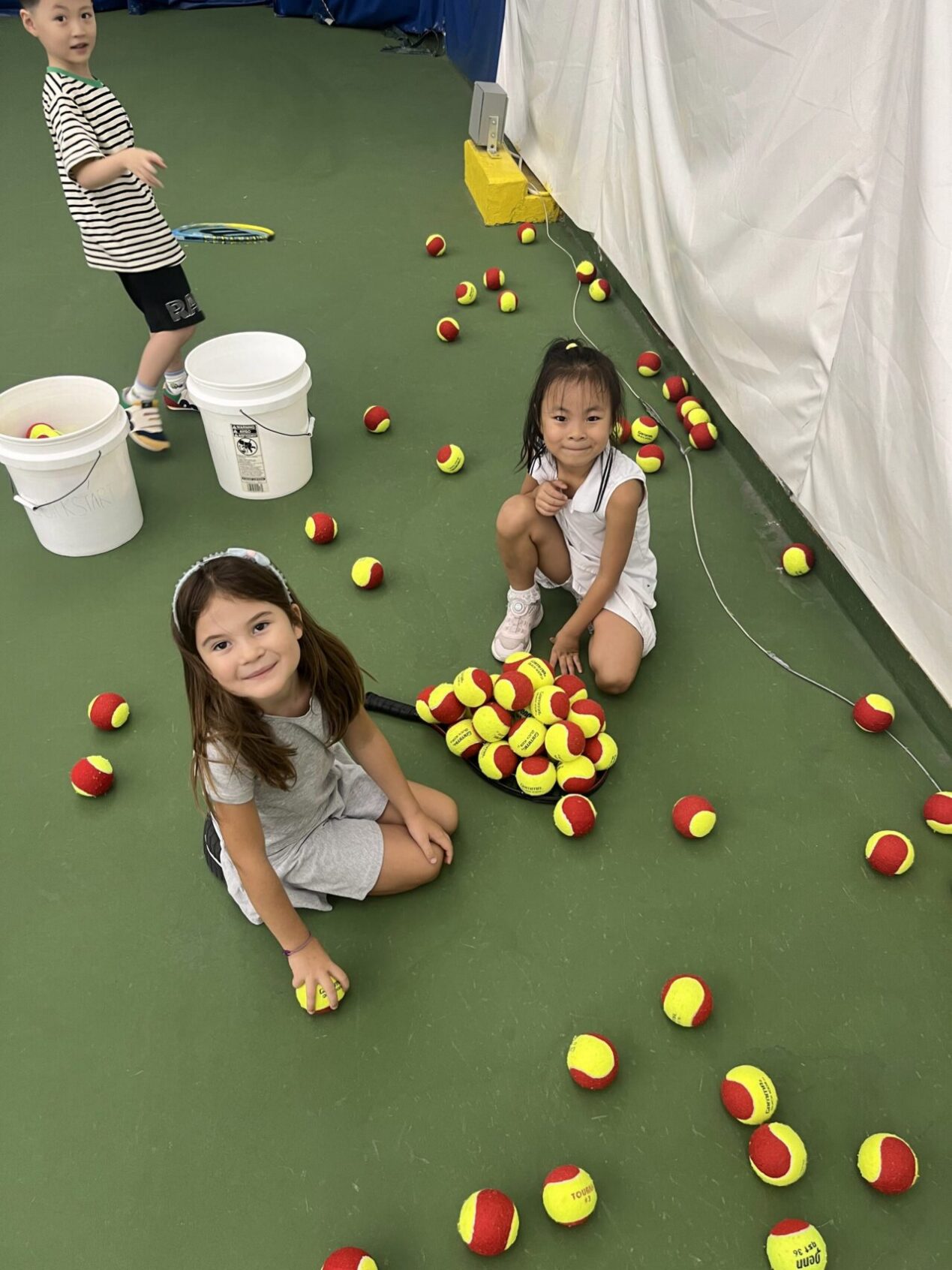 Two girls and a boy picking up tennis balls at an indoor red ball tennis camp in New York.