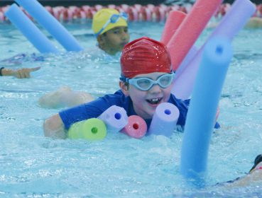 A smiling child with several pool noodles and goggles swimming in a pool at camp.