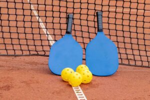 Two pickleball paddles and yellow pickleballs on a clay court.