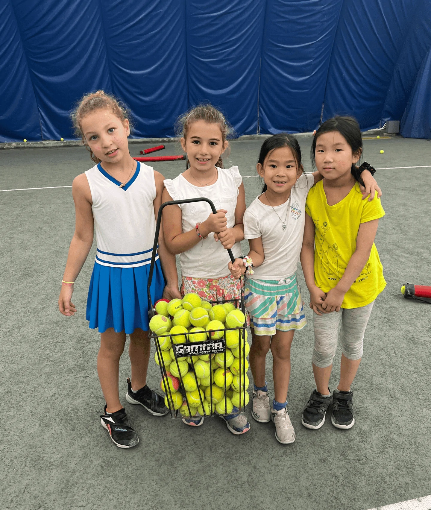 Four girls holding a hopper of tennis balls on an indoor court.